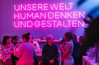 „Unsere Welt human denken und gestalten“: Über den Humanistischen Verband Berlin-Brandenburg Im Haus des HUMANISMUS in der Potsdamer Straße in Berlin sind Leuchtbuchstaben an der Wand zu sehen. Zu lesen ist in Großbuchstaben: „Unsere Welt human denken und gestalten“. Davor sitzen Menschen an einem Tresen und unterhalten sich.