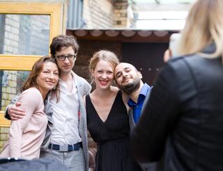 Humanistische Lebensfeiern in Berlin und Brandenburg Vier Menschen in festlicher Kleidung stehen nebeneinander im Freien und werden von einer Frau mit dem Handy fotografiert.