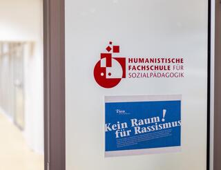 Über die Humanistische Fachschule für Sozialpädagogik in Berlin Auf einer Tür im Inneren des Fachschulgebäudes ist das Schullogo zu sehen. Darunter klebt ein DIN-A4-Blatt auf dem in Weiß auf Blau als Überschrift „Kein Raum für Rassismus!“ plus die Erklärung dazu zu lesen sind.