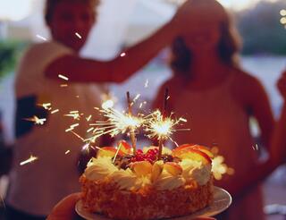 Spenden satt Geschenke Auf dem Bild ist eine kleine Geburtstagstorte zu sehen und zwei Menschen im Hintergrund. Die eine Person hält der anderen die Augen zu.