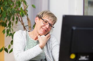 Kontakt- und Beratungsangebote Zu sehen ist eine Frau mit einer Brille und kurzen, blonden Haaren, die mit ihrer Schulter ein weißes Telefon an ihr Ohr drückt und auf einen Computerbildschirm vor sich schaut. Sie lächelt leicht.