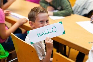Stellenangebote des Humanistischen Verbandes Berlin-Brandenburg Ein Junge sitzt am Tisch und guckt in Richtung der Person, die dieses Bild hier betrachtet. Er hält ein selbst gemachtes Schild hoch, auf dem steht in grünen Großbuchstaben: Hallo.