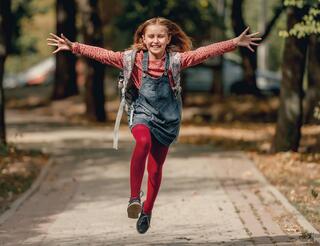 Freie Humanistische Grundschule in Berlin-Pankow Ein Mädchen mit Schulrucksack springt mit ausgestreckten Armen einen Gehweg entlang.