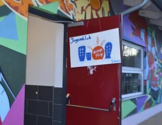Jugendfreizeiteinrichtung Orange Flip Eingang Die Eingangstür des Gebäudes der Jugendfreizeiteinrichtung Orange Flip steht offen. An der Tür hängt ein selbst gemachtes Schild, auf dem in Blau „Jugendclub“ steht, darunter sind drei blaue Hochhäuser, zwischen dem zweiten und dritten ein runder Kreis, in dem in Weiß „Orange Flip“ zu lesen ist. In der Ecke rechts unten ist das Logo des Humanistischen Verbandes Berlin-Brandenburg.