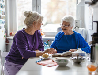 Pflegestützpunkt Neukölln Angehörige Zu sehen sind zwei Frauen, die in einer kleinen Küche an einem weißen Tisch sitzen. Die Frau rechts ist sehr alt und lächelt die andere an. Vor ihr liegt ein Buch, das sie sich zusammen anzuschauen scheinen. Die jüngere Frauch links von ihr lächelt zurück.