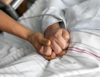 Dong Ban Ja Hospizdienst Zu sehen sind zwei Hände, die sich drücken. Eine Hand gehört zu einer Person, die in einem Bett liegt und in der ein Schlauch steckt.