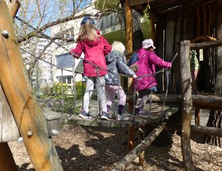 Kinder der Kita Stahlheimer Straße besuchen die Kita Thomas-Mann-Straße: Auf der Hängebrücke Drei Mädchen laufen über eine Hängebrücke im Kitagarten.