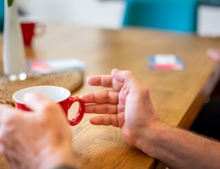 KIS: Kontakt- und Informationsstelle für Selbsthilfe in Berlin-Pankow Eine linke Hand hält eine auf einem Tisch stehende Tasse, die rechte ist horizontal auf der Tischplatte.