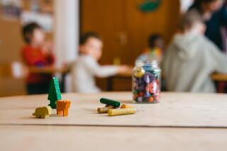 Humanistische Kita Grabbeallee in Berlin-Pankow Auf einem Tisch stehen lHolzfiguren und ein Glas mit Fettstiften, einige Stifte liegen vor dem Glas. Im Hintergrund sind unscharf fünf Kinder zu sehen.