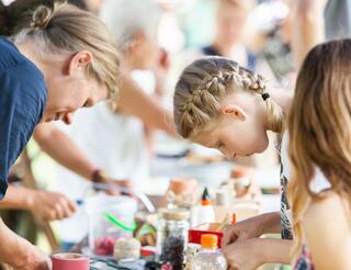 Pressefotos Engagement Eine Frau links und zwei Mädchen rechts basteln etwas an einem Tisch im Freien bei sommerlichem Wetter.