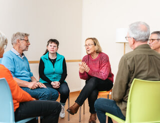 Kontaktstelle PflegeEngagement: Unterstützung für pflegende Angehörige Zu sehen ist eine Gesprächsrunde von sechs Personen, die auf Stühlen in einem Kreis sitzen. Im Zentrum sitzen zwei Frauen. Die Frau rechts trägt einen roten Pulli und schein gerade zu sprechen. Sie hat den Mund leicht geöffnet und ist nach vorne gebeugt.