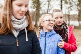 Junge Humanist*innen Berlin-Brandenburg Drei Mädchen in warmen Jacken sind vom Bauch aufwärts zu sehen, im Hintergrund Bäume vor einem Gewässer.