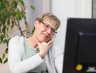 Kontakt- und Beratungsangebote Zu sehen ist eine Frau mit einer Brille und kurzen, blonden Haaren, die mit ihrer Schulter ein weißes Telefon an ihr Ohr drückt und auf einen Computerbildschirm vor sich schaut. Sie lächelt leicht.