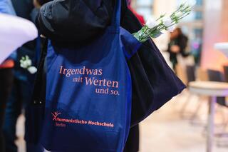 Humanistischer Verband Berlin-Brandenburg: Humanistische Hochschule Berlin Auf einem dunkelblauen Beutel, den eine Person über ihrer linken Schulter trägt, steht: „Irgendwas mit Werten uns so.“ Das Logo der Humanistischen Hochschule Berlin ist unten auf dem Beutel in Orange zu sehen.